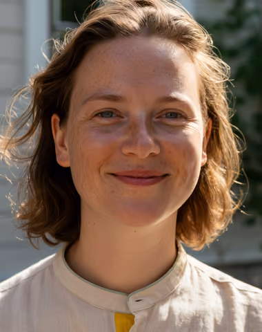 Smiling young woman with short wavy hair and beige shirt outdoors.