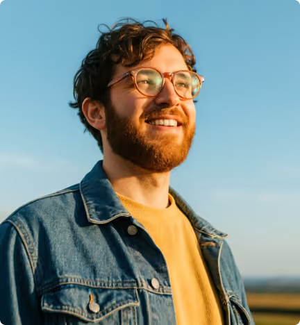 Smiling man with glasses and a beard wearing a denim jacket outdoors under clear blue sky.