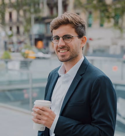 Young man in glasses and a suit jacket holding a takeaway coffee cup outdoors in an urban setting.