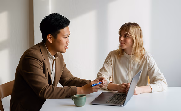 Man and woman sitting at table with laptop and coffee cup, engaged in conversation.