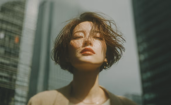 Woman with short hair and closed eyes standing outdoors in front of tall blurred buildings.