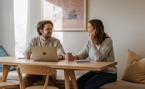 Two people smiling and talking at a table with a laptop and coffee cups in a cozy room.