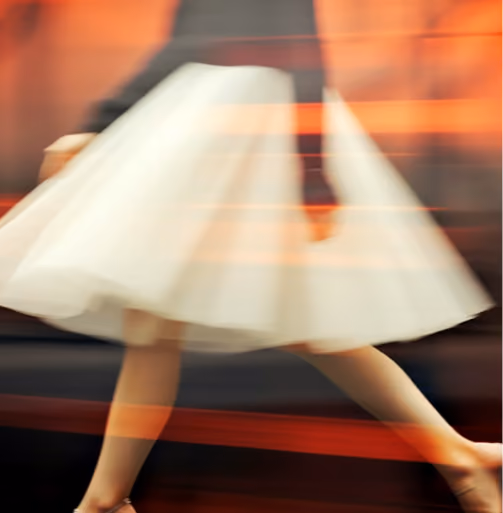 Blurred image of person in white skirt walking, creating motion effect with orange light streaks.