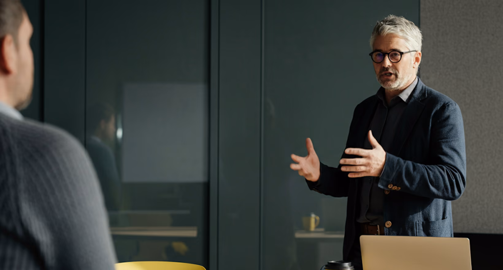 Man with gray hair and glasses speaking and gesturing in a modern office setting.