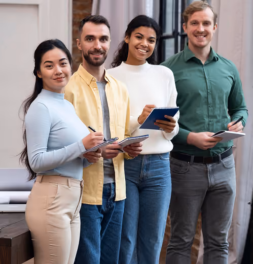 Four smiling young adults standing with notepads and pens in a modern indoor setting.