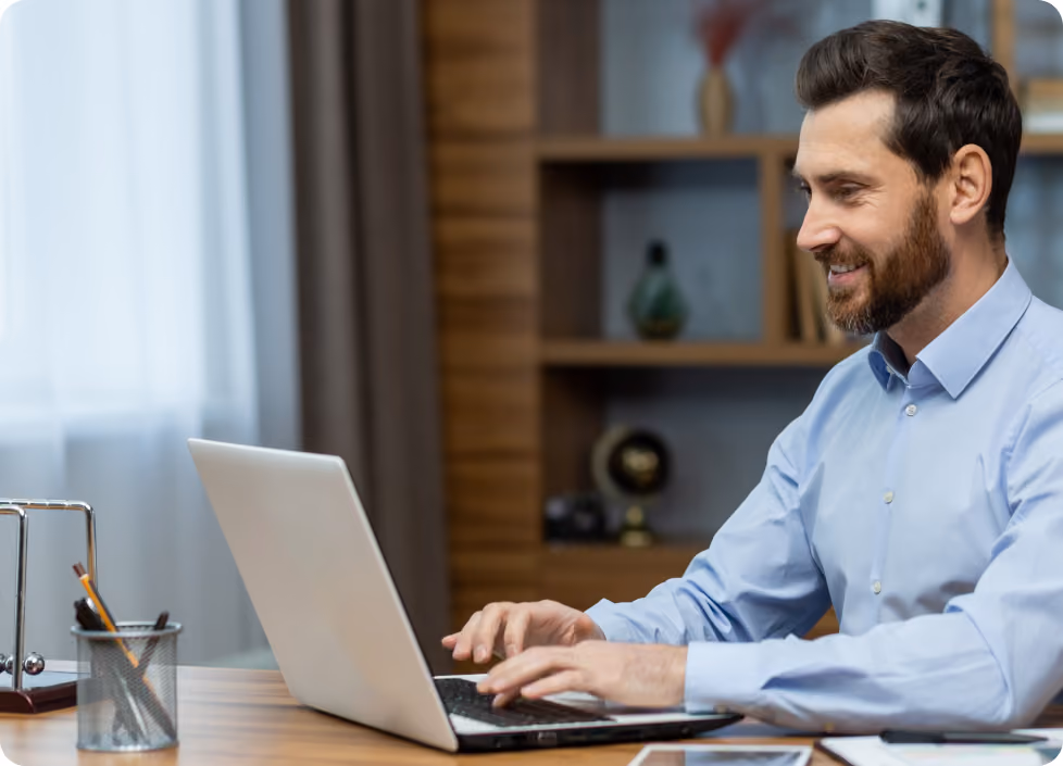 Smiling bearded man in blue shirt typing on laptop at wooden desk in modern office.
