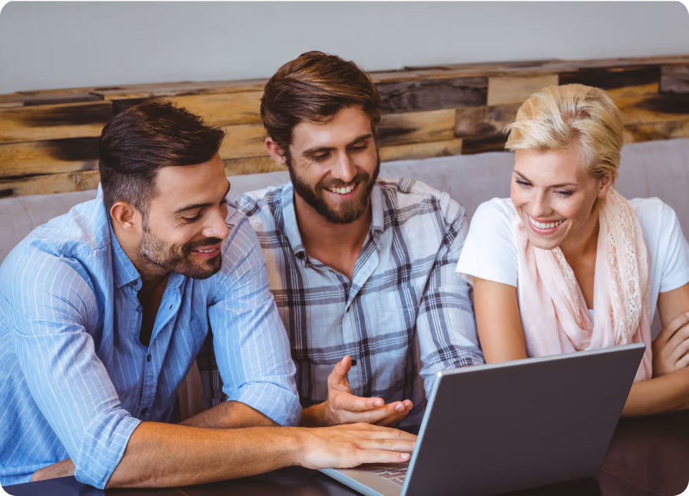 Three smiling adults sitting closely and looking at a laptop on a table indoors.