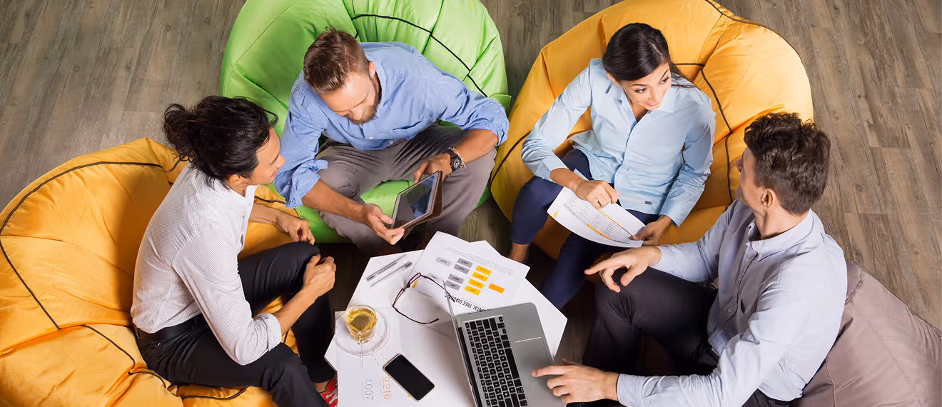 Four professionals discuss documents and digital devices seated on colorful bean bags around a white table.
