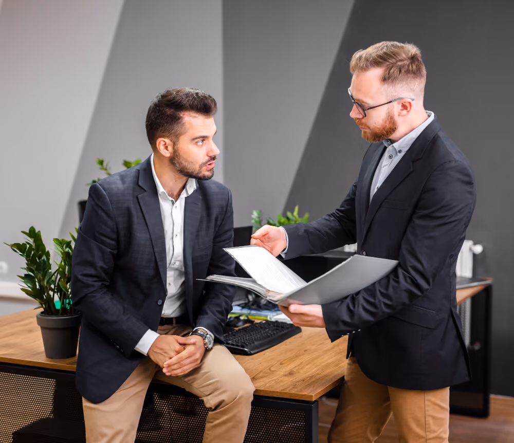 Two men in business attire discussing documents in an office with plants and a desk.