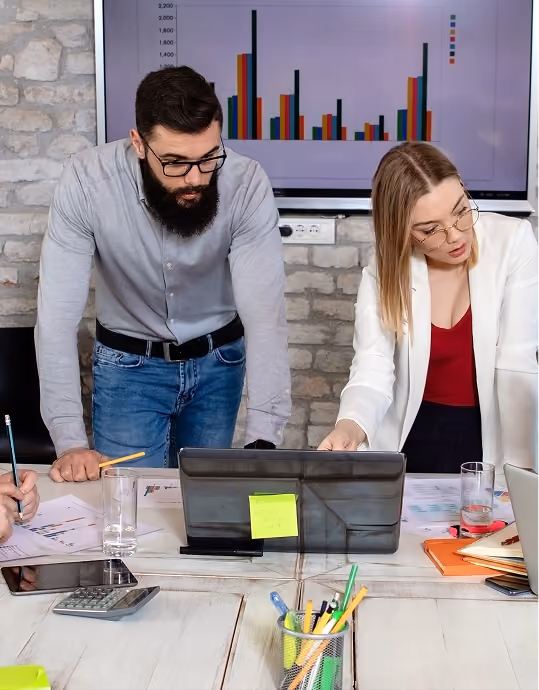 Two professionals review documents at a table with a laptop and a bar chart on a screen behind them.