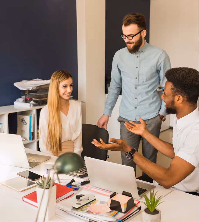 Three colleagues having a friendly discussion around laptops in a modern office.
