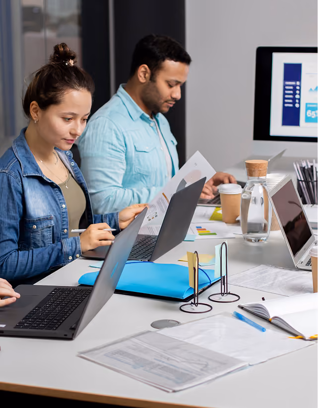 Two people working at a desk with laptops, papers, coffee cups, and a screen showing charts.