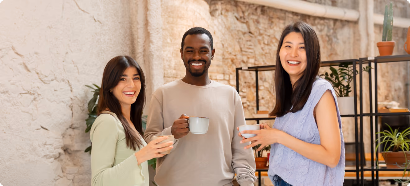 Three diverse adults smiling and holding coffee mugs in a relaxed indoor setting with plants.