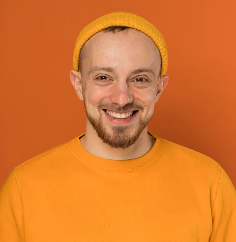 Smiling man with a mustache and beard wearing a yellow beanie and yellow sweater against an orange background.