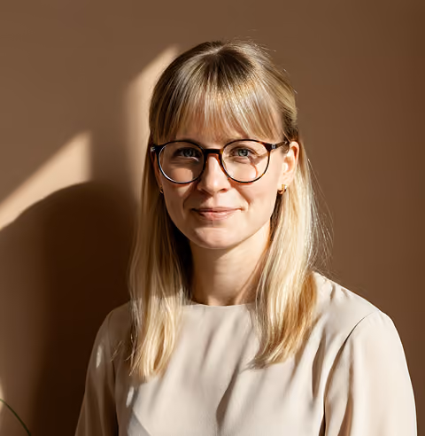 Woman with blonde hair wearing round glasses and a beige top against a brown background.