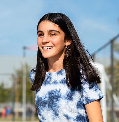 Smiling young woman with long dark hair wearing a blue and white tie-dye shirt outdoors.