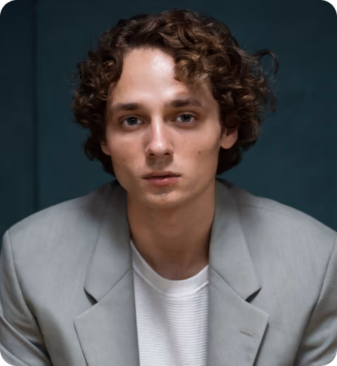 Young man with curly hair wearing a gray blazer and white shirt against dark background.