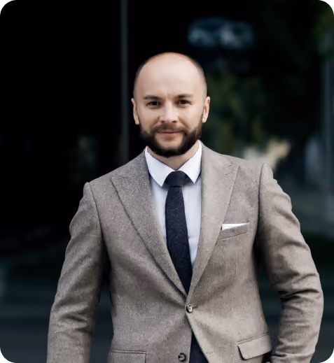 Man with a beard wearing a brown suit jacket, white shirt, and black tie standing outdoors.