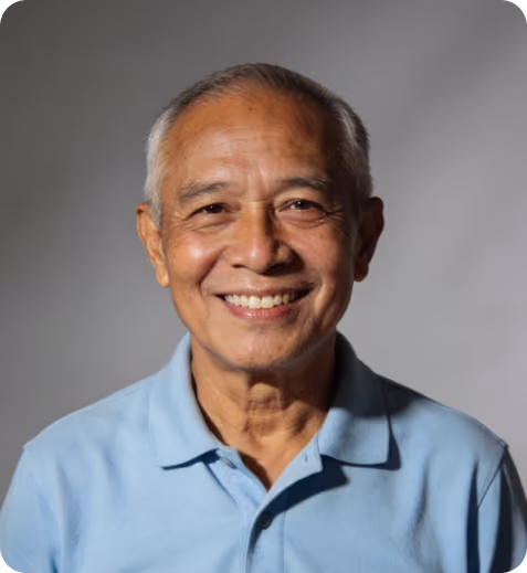 Smiling elderly man wearing a light blue polo shirt against a plain gray background.