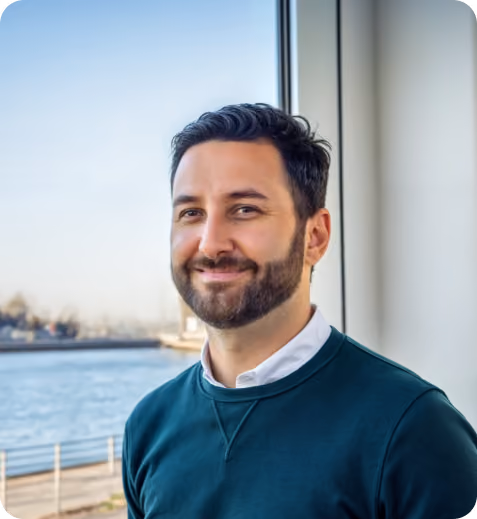 Smiling man with dark hair and beard wearing a teal sweater standing by a window overlooking water.