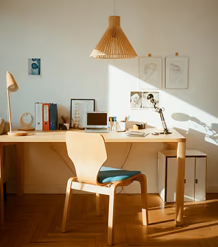Wooden desk and chair in a sunlit room with a laptop, lamp, and office supplies.