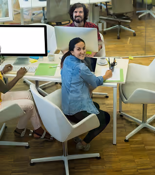 Three coworkers sitting at a table in an office, smiling and working on laptops and documents.
