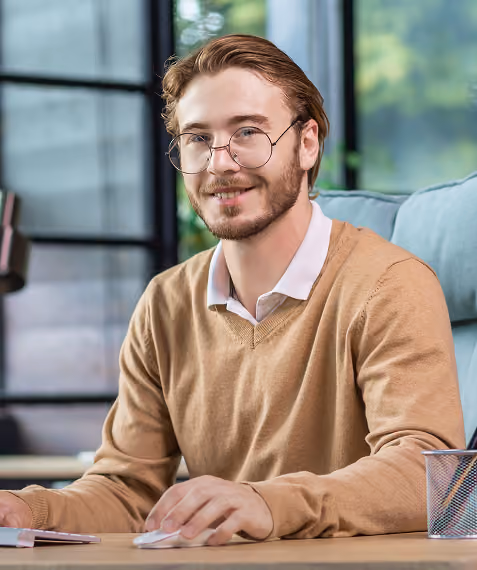 Man with glasses and brown sweater sitting at desk using a computer mouse, smiling at camera.
