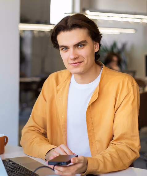 Young man with dark hair wearing mustard shirt and white tee, holding phone, sitting at desk with laptop.