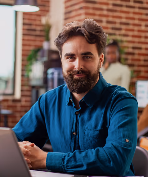 Bearded man in a blue shirt smiling at laptop in a cozy office with brick wall background.