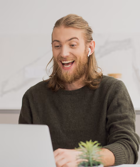 Smiling man with long hair and beard wearing wireless earbuds looks at laptop screen indoors.