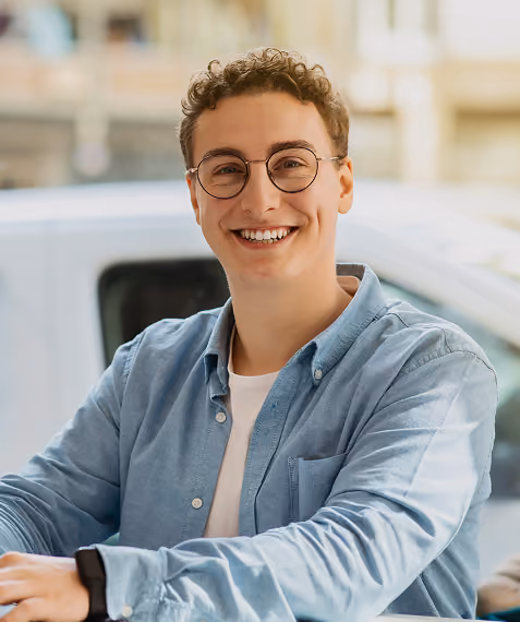 Smiling young man with curly hair and glasses wearing a light blue shirt indoors.