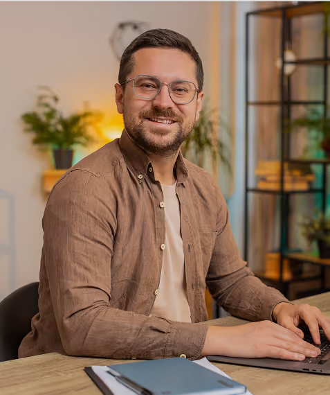 Man with glasses smiling while typing on a laptop at a desk with a notebook and pen.