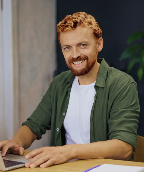 Smiling man with red hair and beard wearing green shirt working on a laptop at a desk.