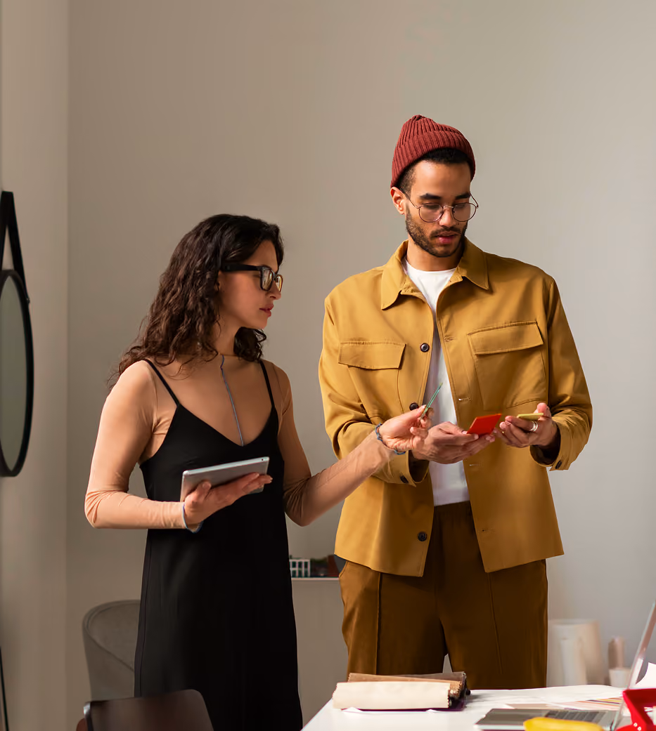 Woman showing color swatches to man wearing mustard jacket and red beanie in office.