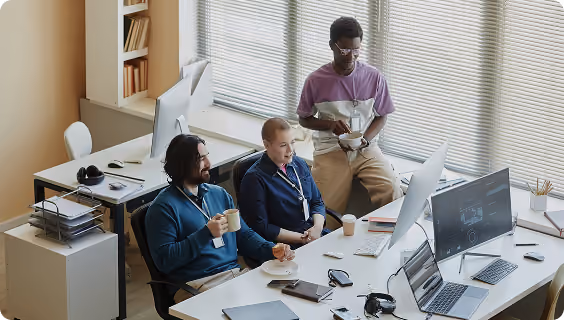 Three coworkers in an office with computers, two seated talking and one standing holding a coffee cup.