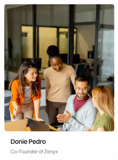 Four colleagues smiling and discussing work around a laptop in an office setting.