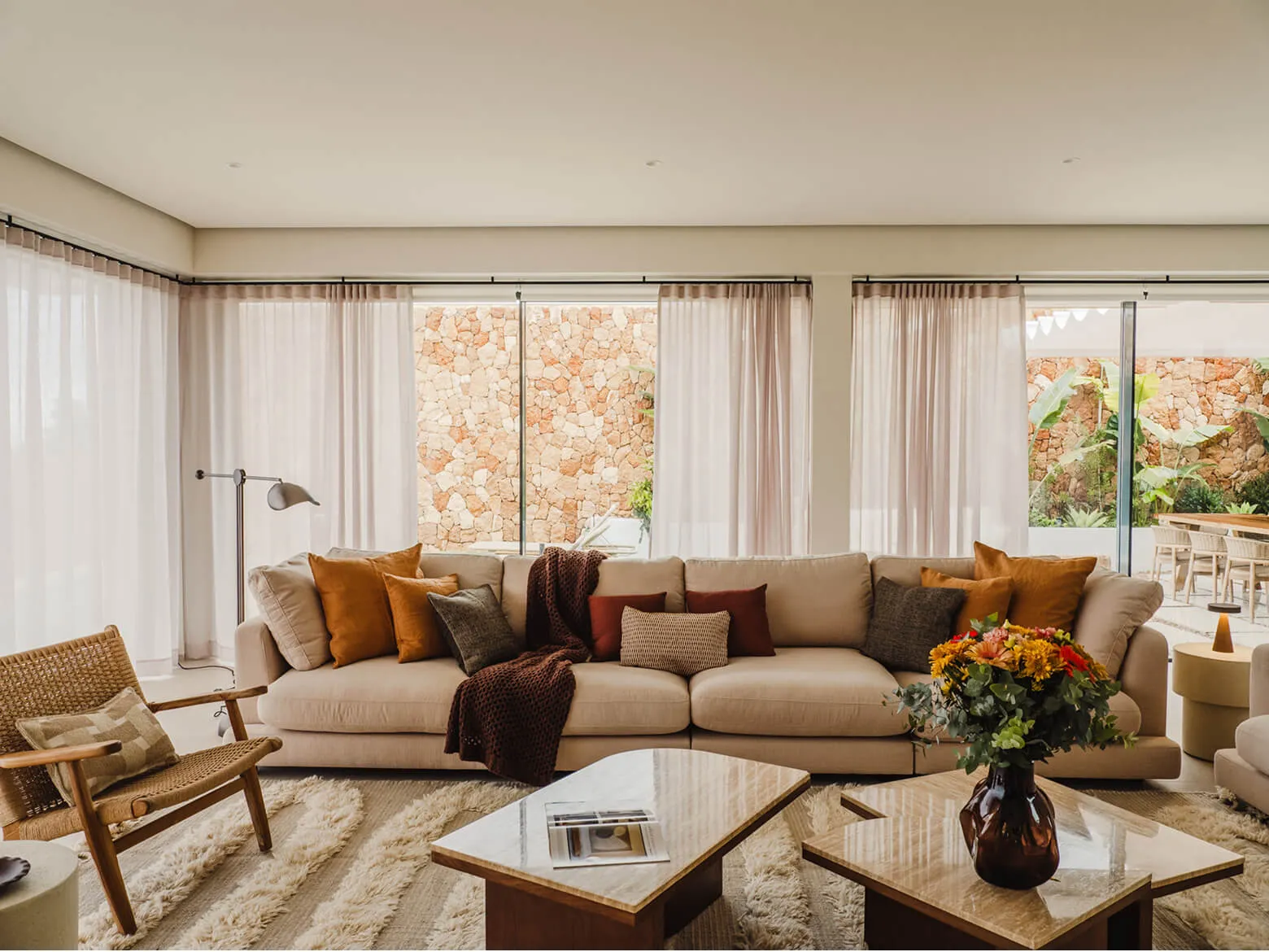 High-end interior design and styling of a bright villa living room in Ibiza, showcasing a neutral linen sofa with terracotta cushions and natural stone walls visible through sheer drapes.