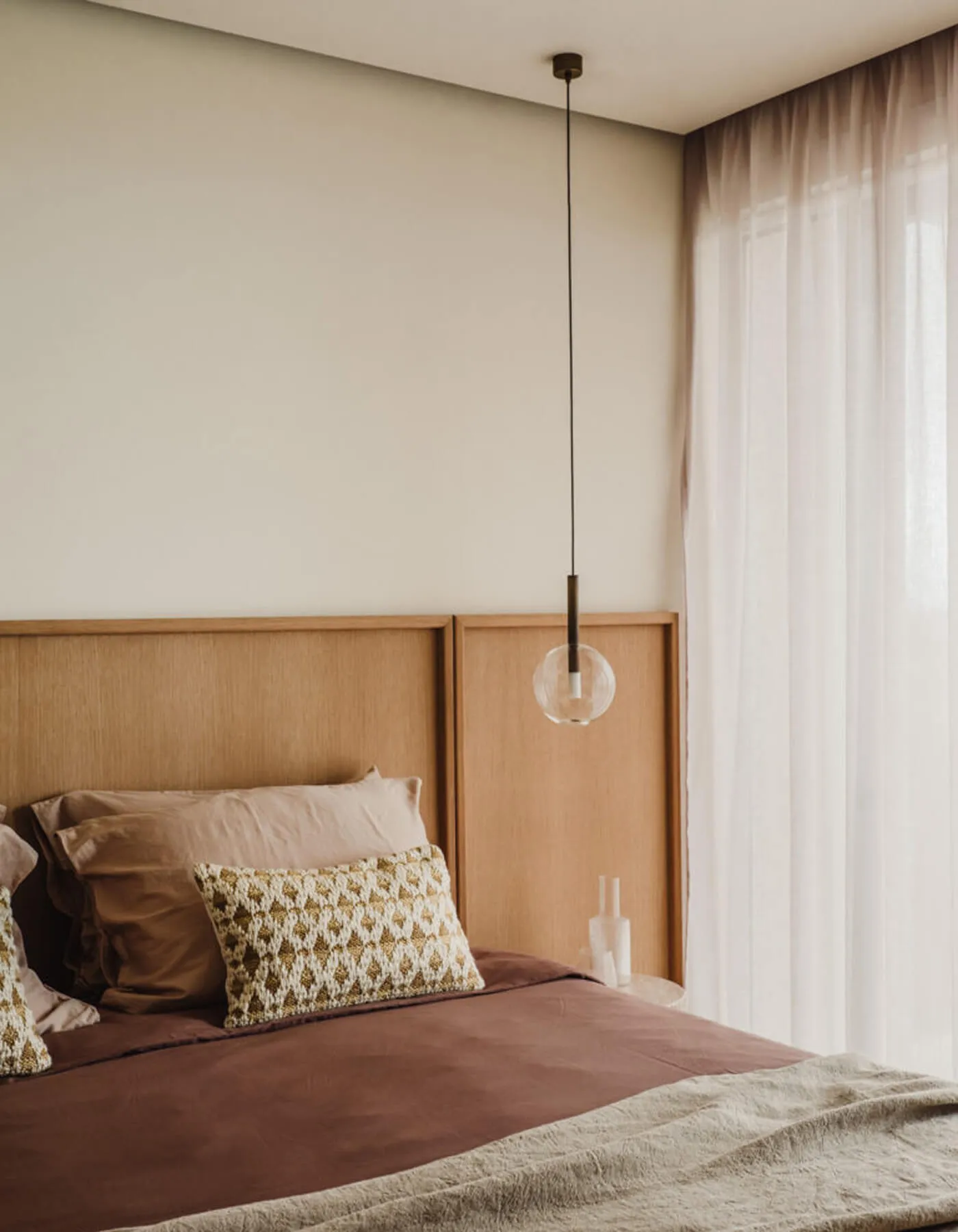 High-end residential interior design in Ibiza, featuring a sleek warm wood panel headboard, plum-toned premium linens, and a floating glass sphere pendant light.
