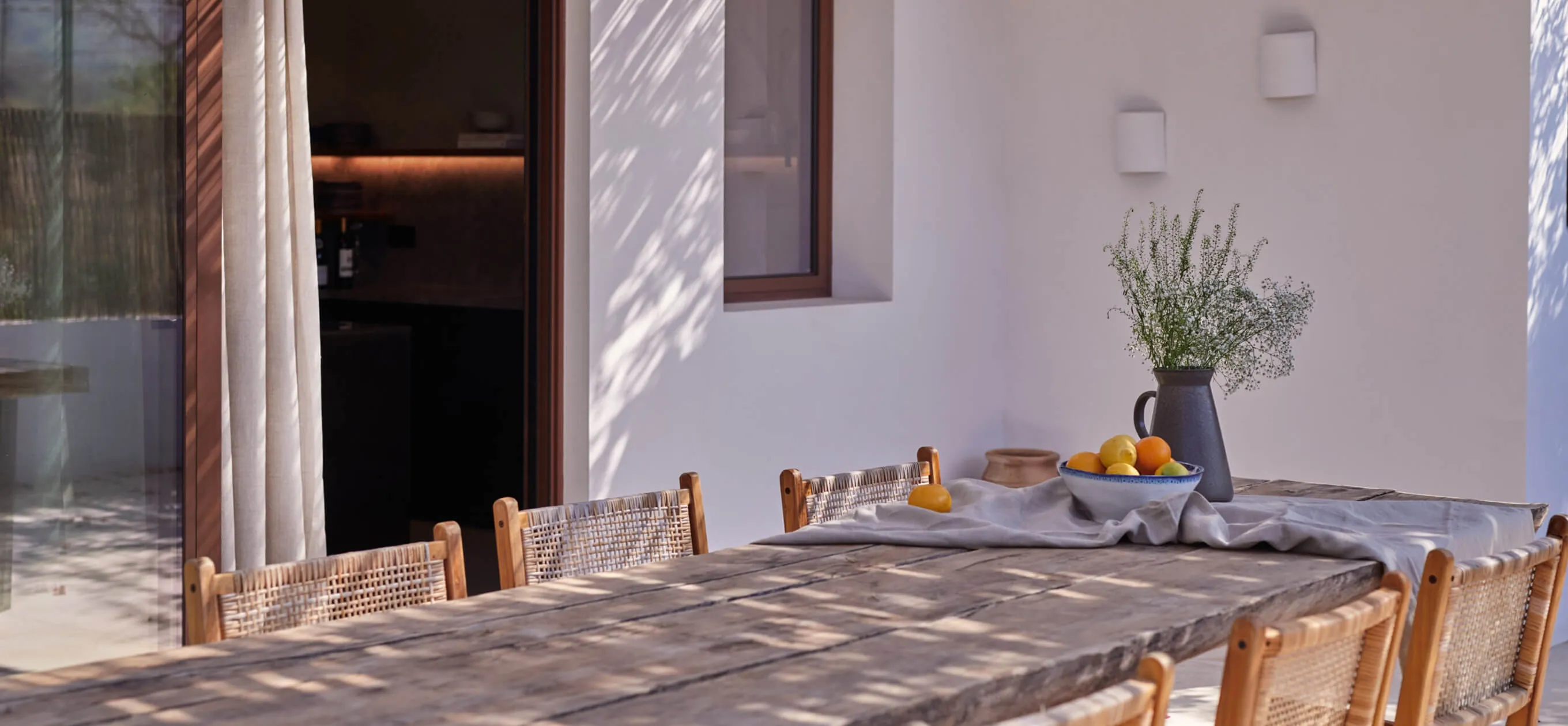 Luxury outdoor dining area at the Sa Caleta project in Ibiza, featuring a rustic wooden table, woven chairs, and soft pergola shadows.