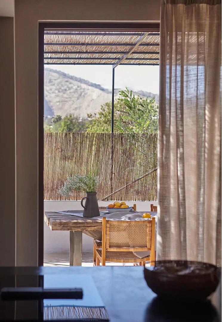 View from a modern kitchen looking out to a shaded rustic terrace at Sa Caleta, framed by elegant sheer linen curtains in Ibiza.