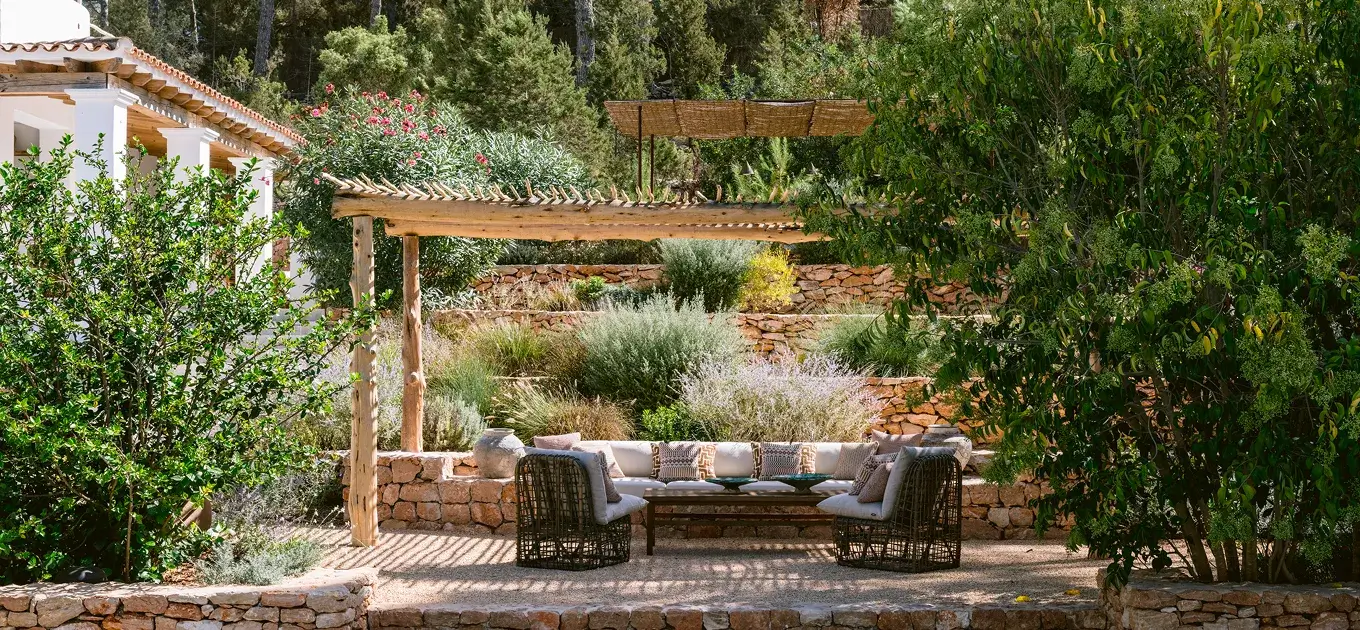Luxury outdoor lounge and terrace interior design at Villa Benimussa in Ibiza, featuring a rustic wooden pergola, traditional stone walls, and premium terrace furniture surrounded by Mediterranean greenery.