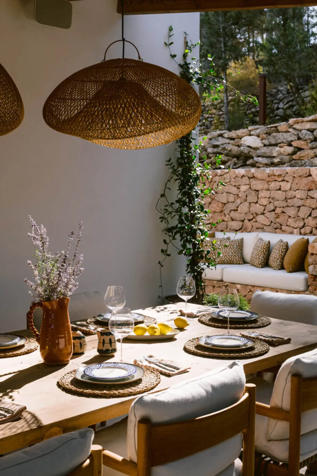 High-end outdoor dining terrace interior design at Villa Benimussa, Ibiza, with a rustic wood table, woven lamps, and a dry stone wall backdrop.