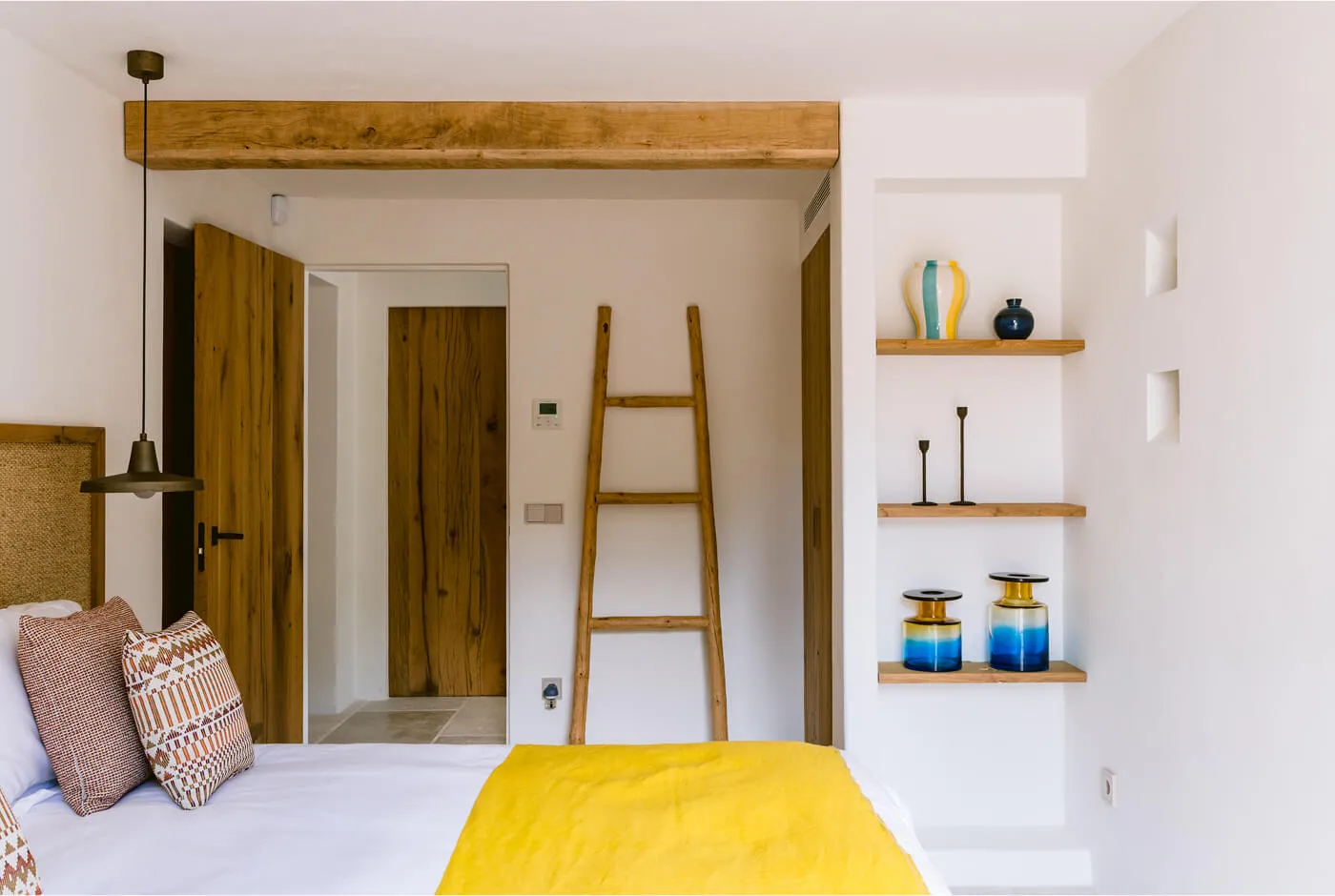 Vibrant bedroom interior architecture at Villa Benimussa, featuring a bright yellow blanket, an exposed wooden ceiling beam, and built-in display shelves.
