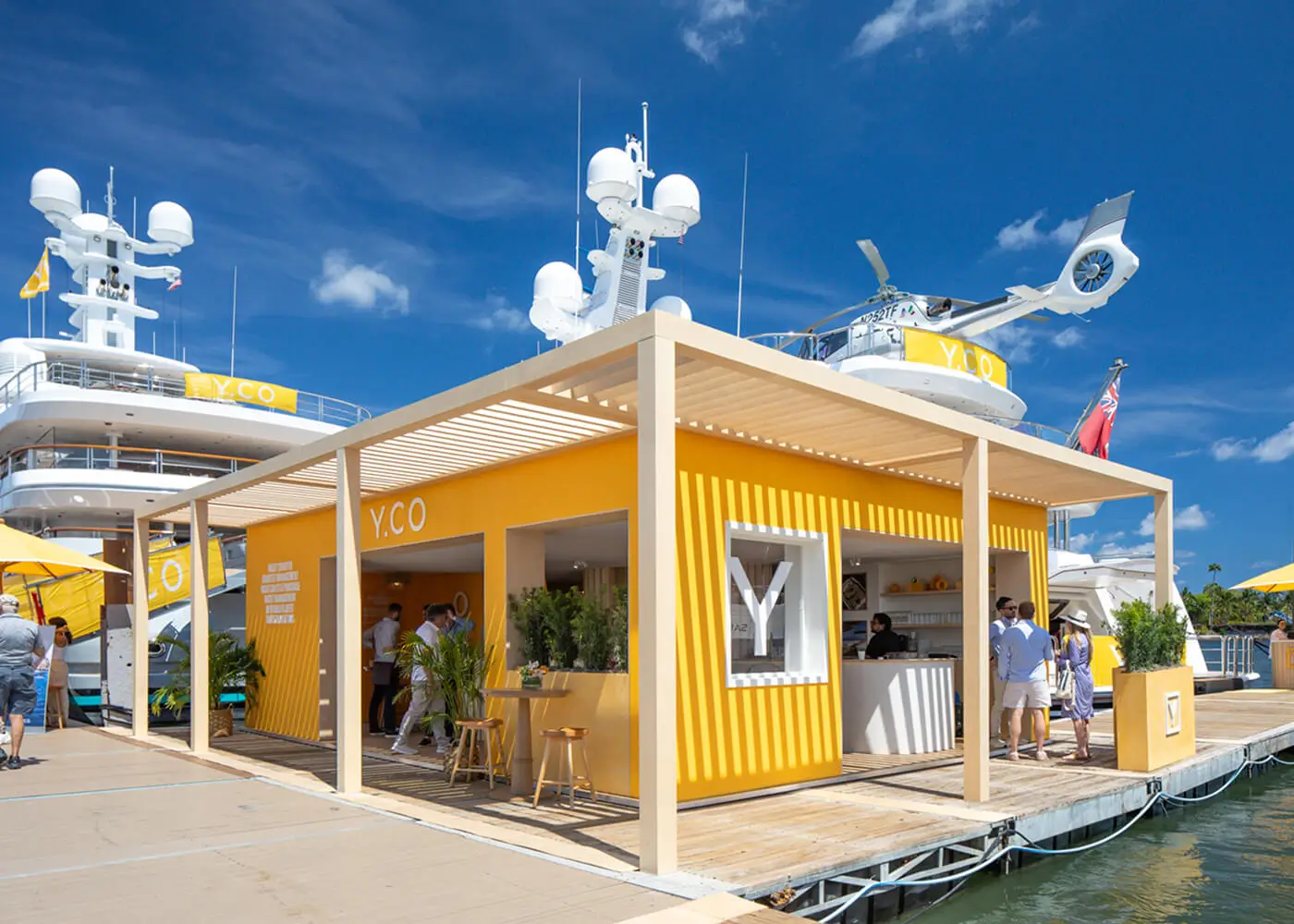 External wide perspective of the yellow contemporary beach club-inspired Y.CO Clubhouse pavilion, built on a floating pontoon surrounded by mega yachts and marine environment scenery.