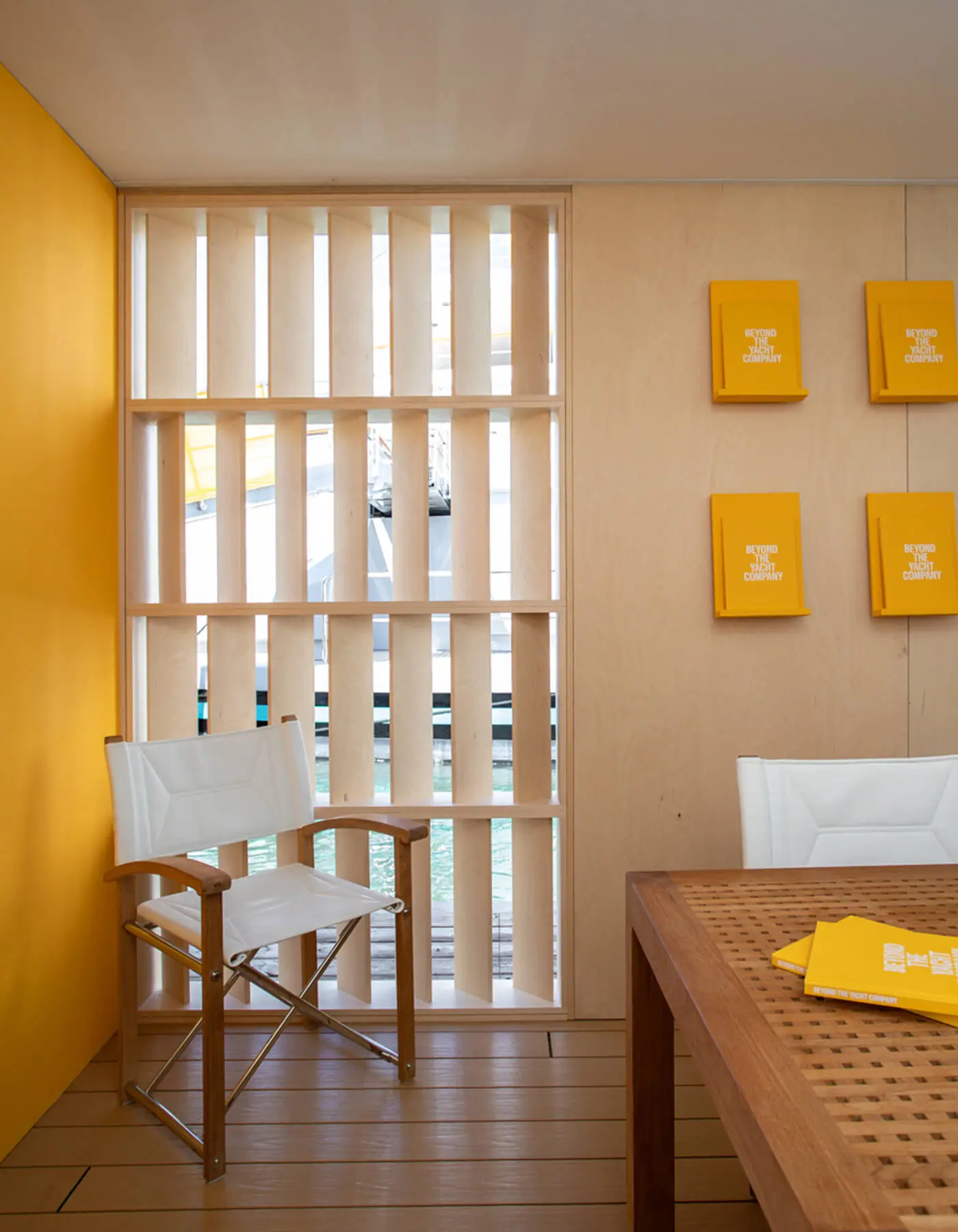 Interior detail of the Y.CO Clubhouse pavilion by Studio Moravová, featuring a white safari director's chair next to custom birch wood vertical louvers and a light timber wall with yellow branded book displays.