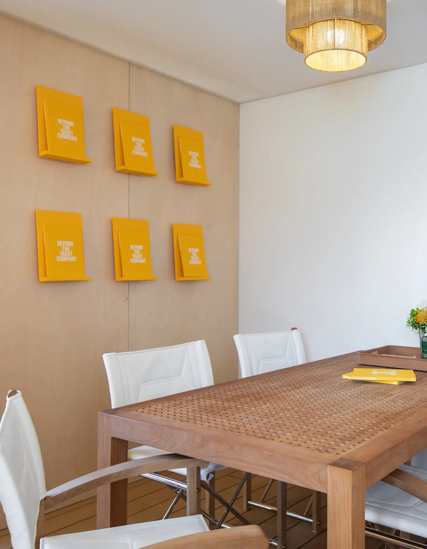 High-end meeting zone inside the Y.CO Clubhouse exhibition pavilion, showcasing a bespoke teak grid table, white leather safari chairs, birch wood joinery walls, and a warm woven rattan pendant light.