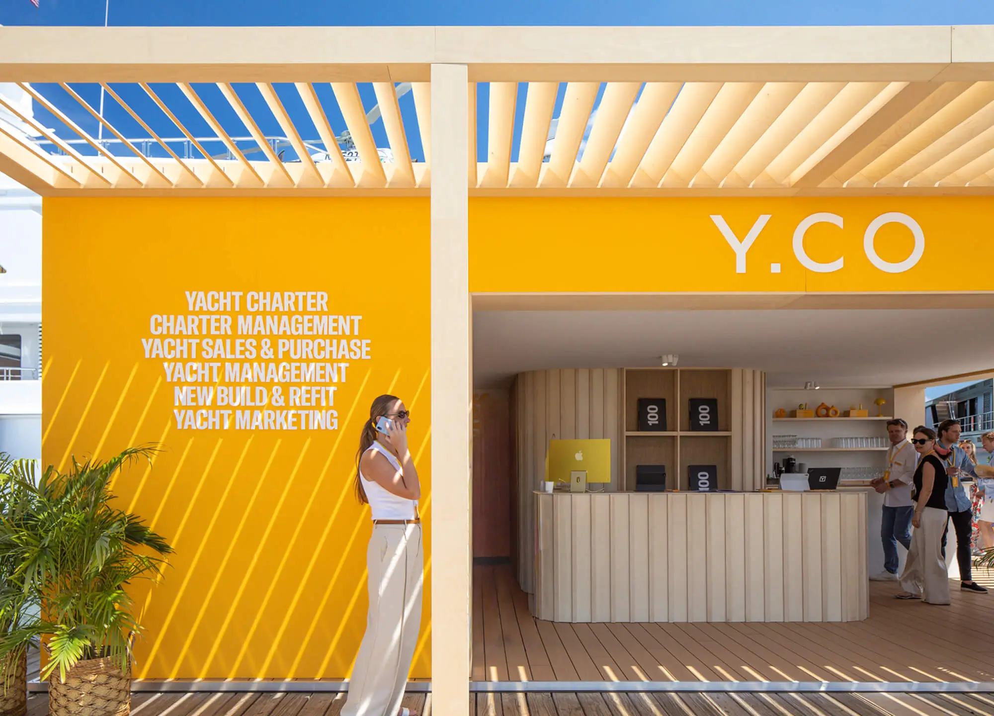 Close-up of the yellow exterior wall of the Y.CO Clubhouse pavilion with white typography listing yacht marketing and management services next to the open fluted wood counter.