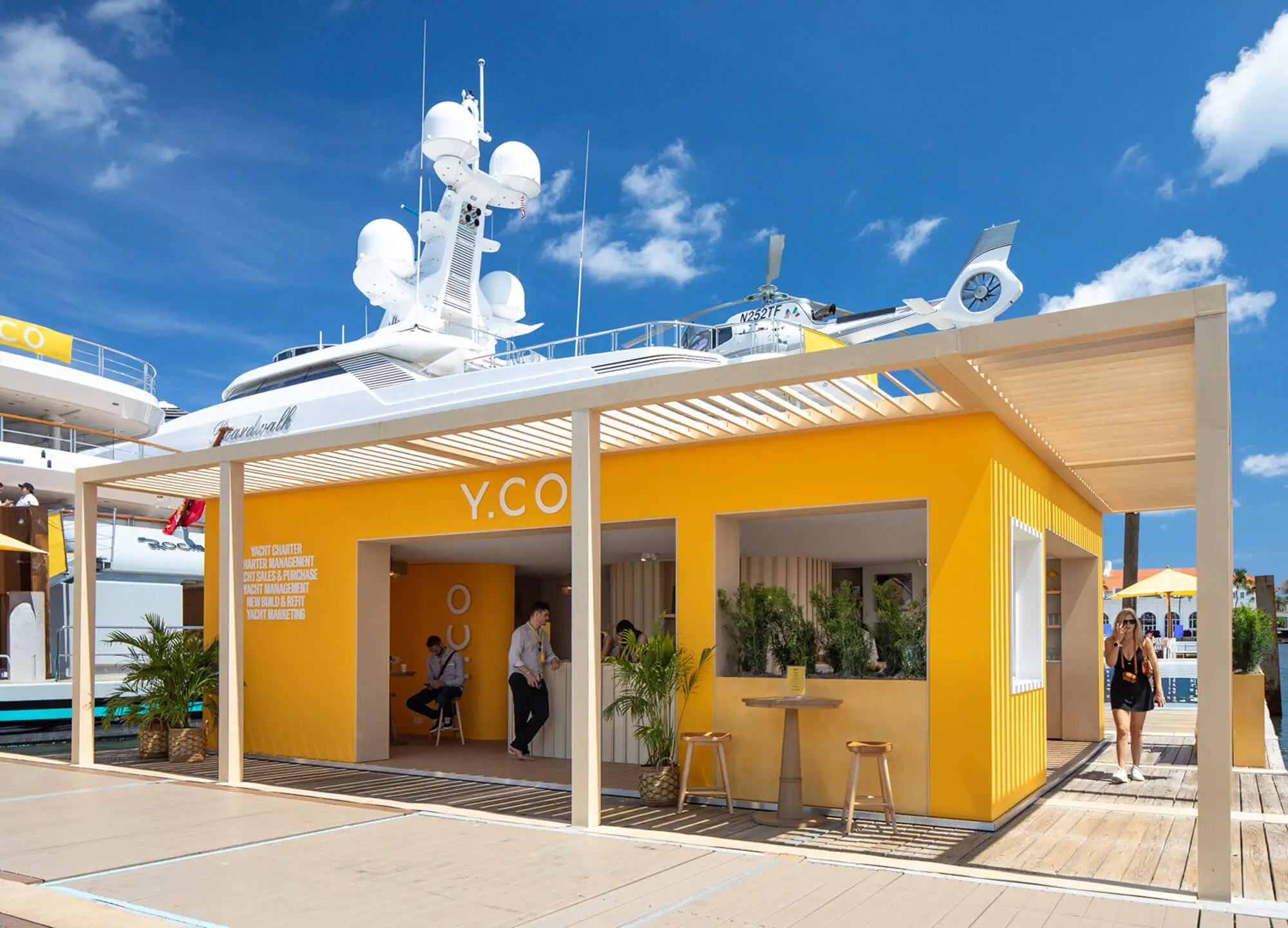 Floating teak deck walkway alongside the modular Y.CO Clubhouse exhibition pavilion, featuring open sightlines, shaded meeting areas, and custom built-in plant containers.