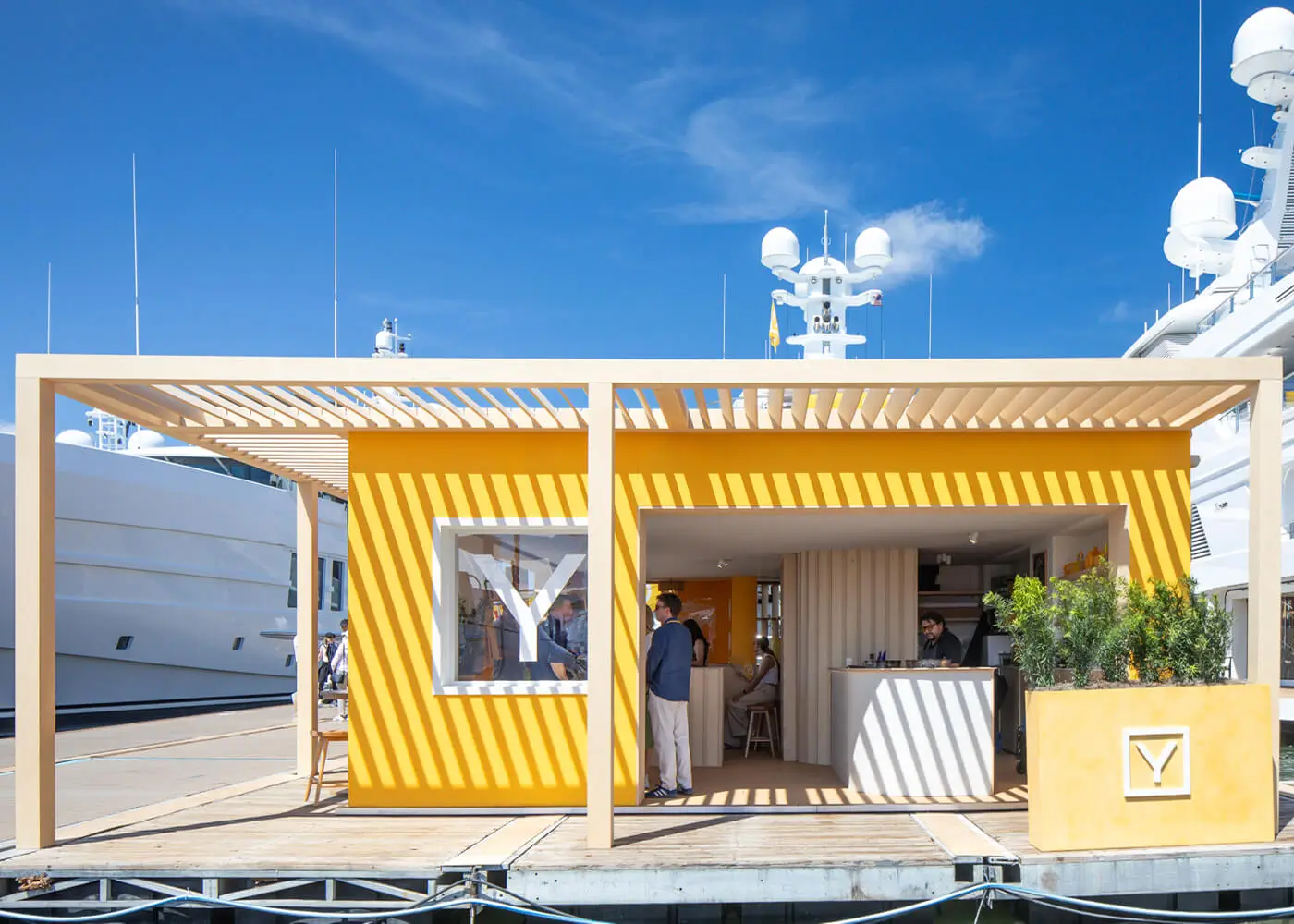 Striking parallel shadows cast across the yellow clay render facade of the Y.CO Clubhouse stand from an overhead timber pergola, highlighting a minimalist white 'Y' brand logo display.