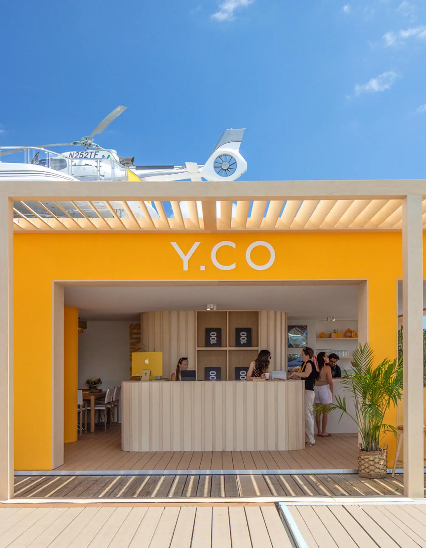 Front view of the Y.CO Clubhouse exhibition stand under a timber pergola ceiling. A fluted birch wood reception desk stands against a bold yellow clay-particle rendered wall, with a white superyacht hull visible overhead.